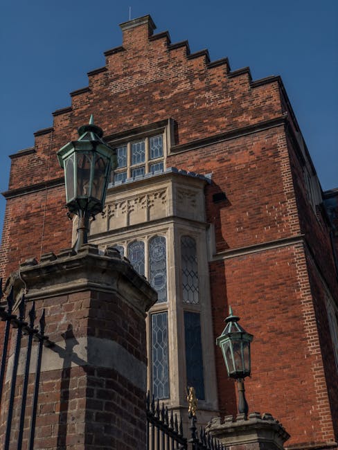 A historic brick building with a symmetrical facade featuring two prominent gables adorned with decorative brickwork, situated behind a black wrought iron fence with stone pillars. The front area incl