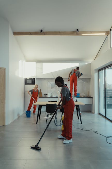 A person wearing protective clothing, including gloves and a suit, is using a vacuum cleaner with a long hose attachment to clean a smooth, polished floor surface. The scene appears to be a commercial