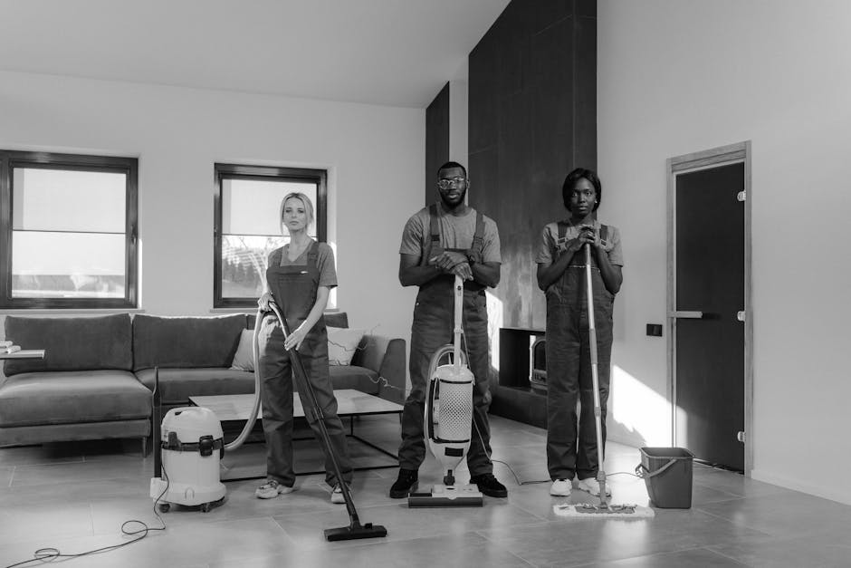 Three professional cleaning staff from Harrow Cleaners are performing surface cleaning and deep cleaning tasks in a modern living room. The room features a light-colored tiled floor, a large L-shaped sofa with a dark fabric cover, and two windows allowing natural light to illuminate the space. The team, dressed in uniform, is using various cleaning tools: one with a vacuum cleaner, another holding a mop, and the third with a cleaning cloth. The room appears thoroughly cleaned and well-maintained, highlighting their focus on maintaining hygiene and cleanliness through meticulous surface and floor cleaning processes. Visible surfaces, including the tiled floor and the sofa, are free of dust and debris, with the vacuum cleaner and mop indicating active sanitation work. Harrow Cleaners provides expert domestic cleaning services, ensuring residential spaces are sanitized and refreshed during end of tenancy cleaning or routine hygiene maintenance.