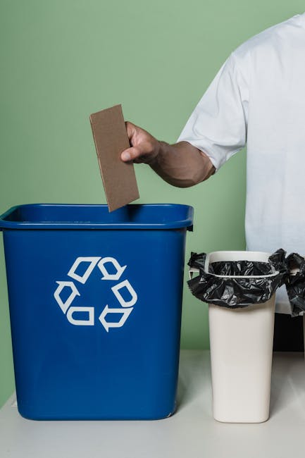 A row of five large outdoor waste bins positioned on a paved surface with some dirt and debris around them, adjacent to a large closed metal storage container. The waste bins include a black paper rec