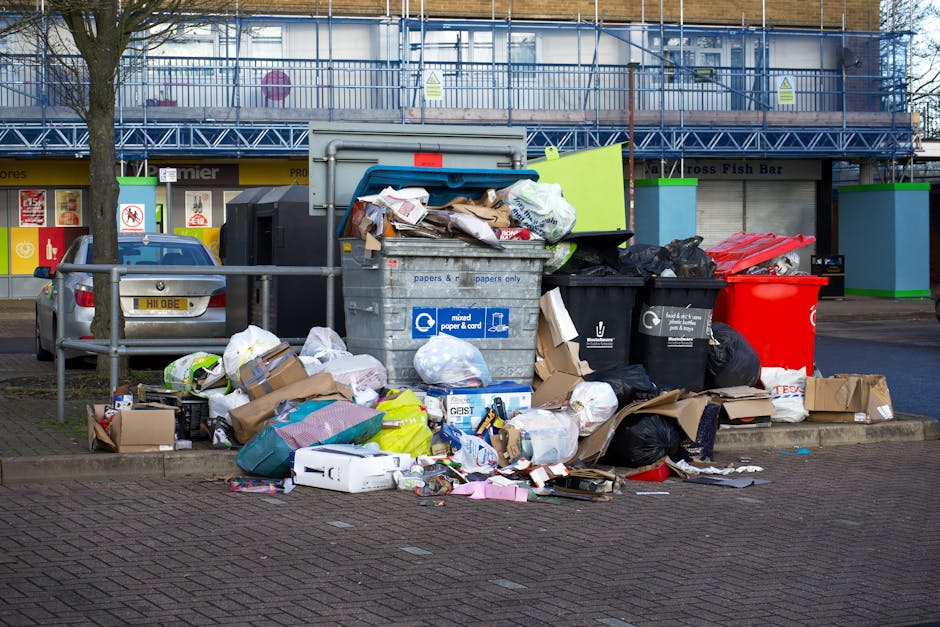 A commercial cleaning trolley and waste bin situated against a tiled beige wall in a cleaning storage area. The trolley holds various cleaning tools such as mops, brooms, spray bottles, and cloths, al